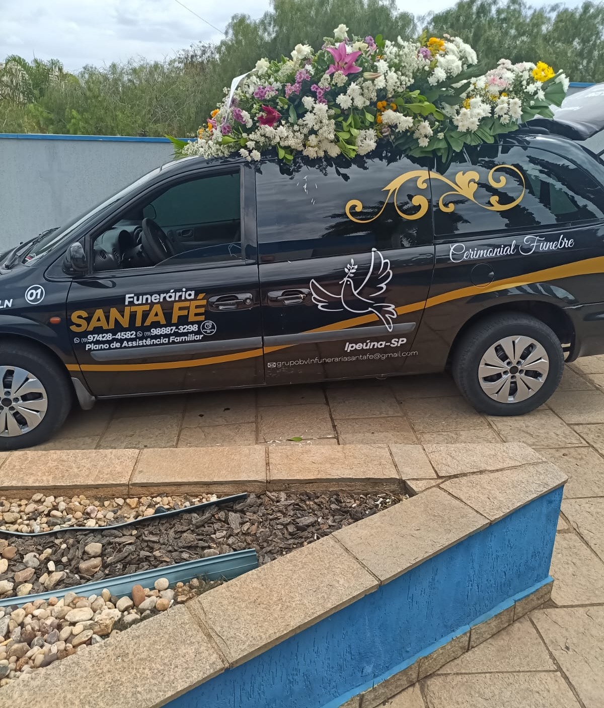 Viatura funerária oficial da Funerária Santa Fé decorada com arranjo floral de flores brancas, lilases e amarelas, exibindo a identidade visual dourada e os con